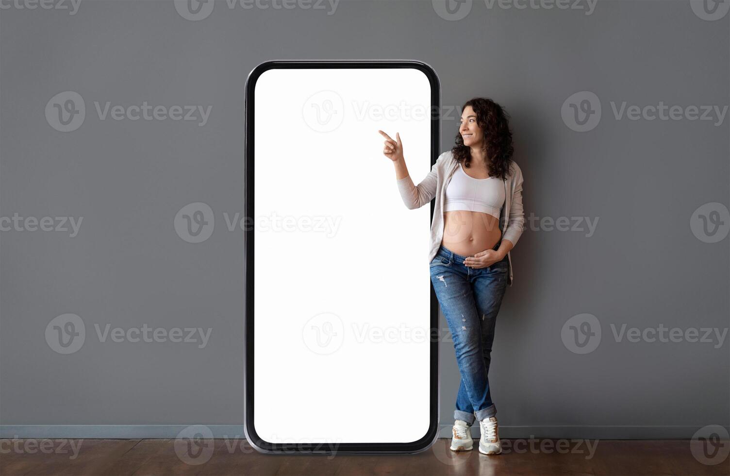 A woman in casual attire stands next to an oversized phone frame, smiling and pointing. She showcases her baby bump, conveying joy and anticipation in a contemporary setting. photo