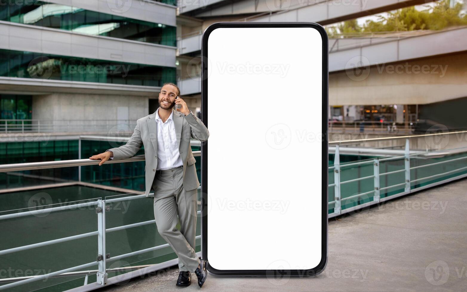 A businessman dressed in a gray suit is casually talking on his smartphone while leaning against a railing, standing by huge phone with blank screen photo