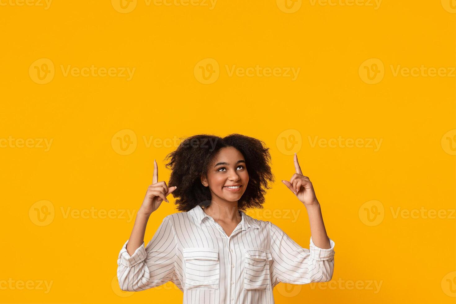 Look There. Happy Afro Girl With Funny Curls Pointing Upwards At Copy Space Above Her Head Over Yellow Background, Vertical Shot photo