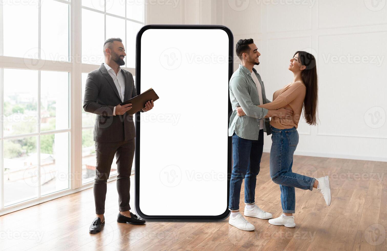 A couple shares a joyful moment in a bright, modern interior. They embrace while a man with a tablet observes, huge phone with blank screen next to them photo