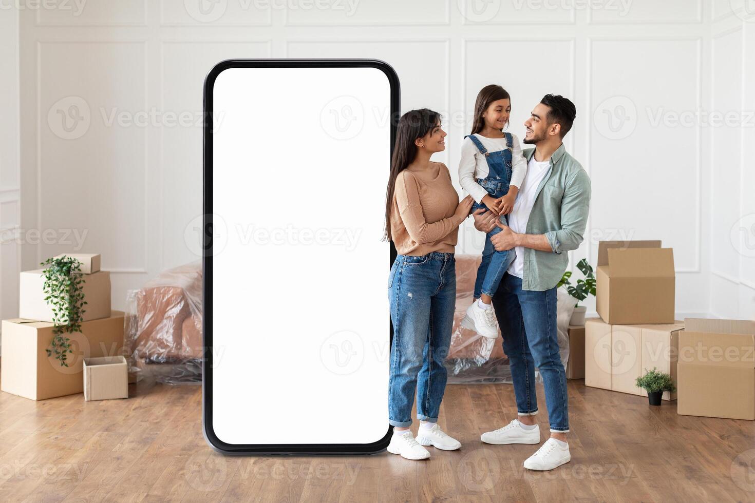 A joyful middle eastern family enjoys their new living space together, standing next to a large empty frame where they may place a TV in their cozy room filled with moving boxes. photo