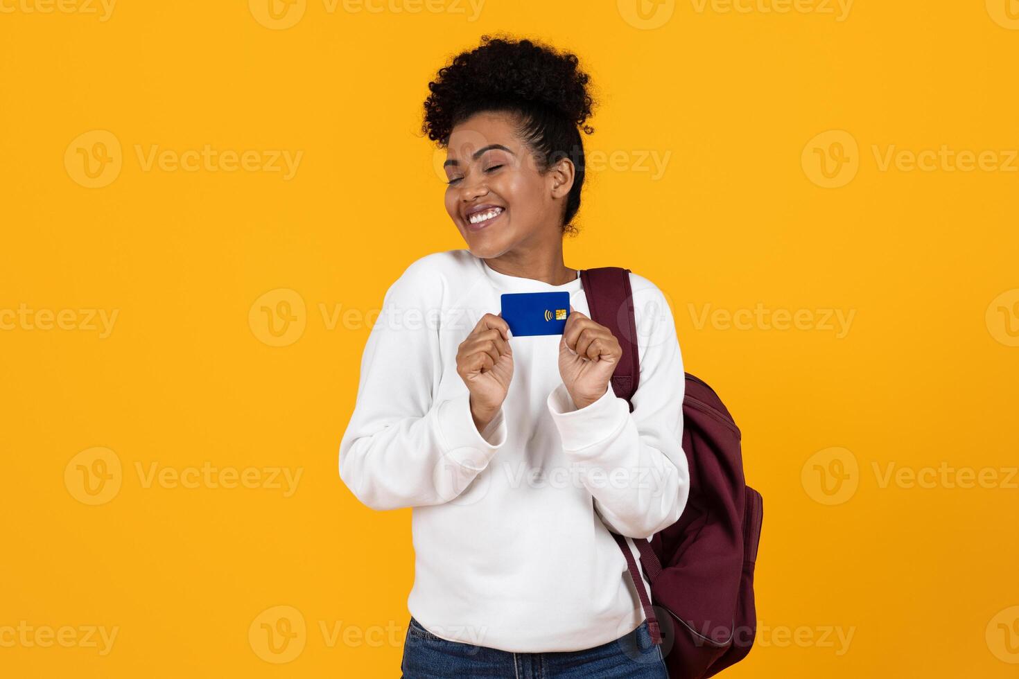 A cheerful young woman in a white sweater and jeans happily displays her credit card while standing against a vibrant yellow backdrop, embodying excitement and financial empowerment. photo
