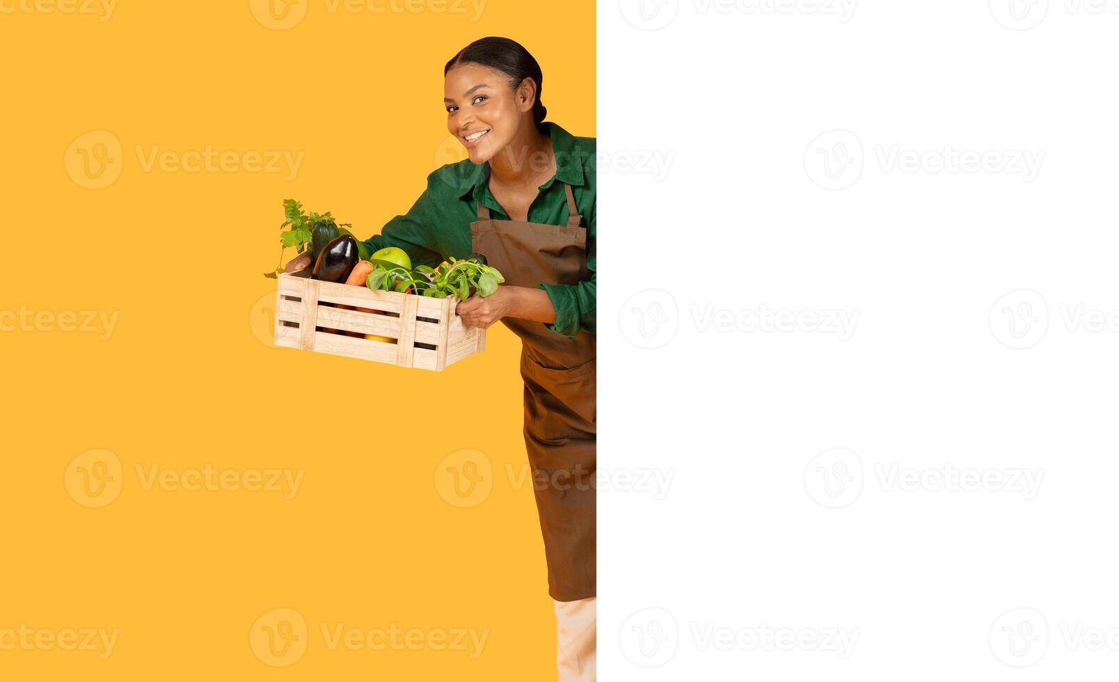 A young woman in an apron joyfully presents a wooden crate filled with fresh vegetables. The vibrant background enhances her cheerful demeanor while showcasing the bounty of fresh produce. photo