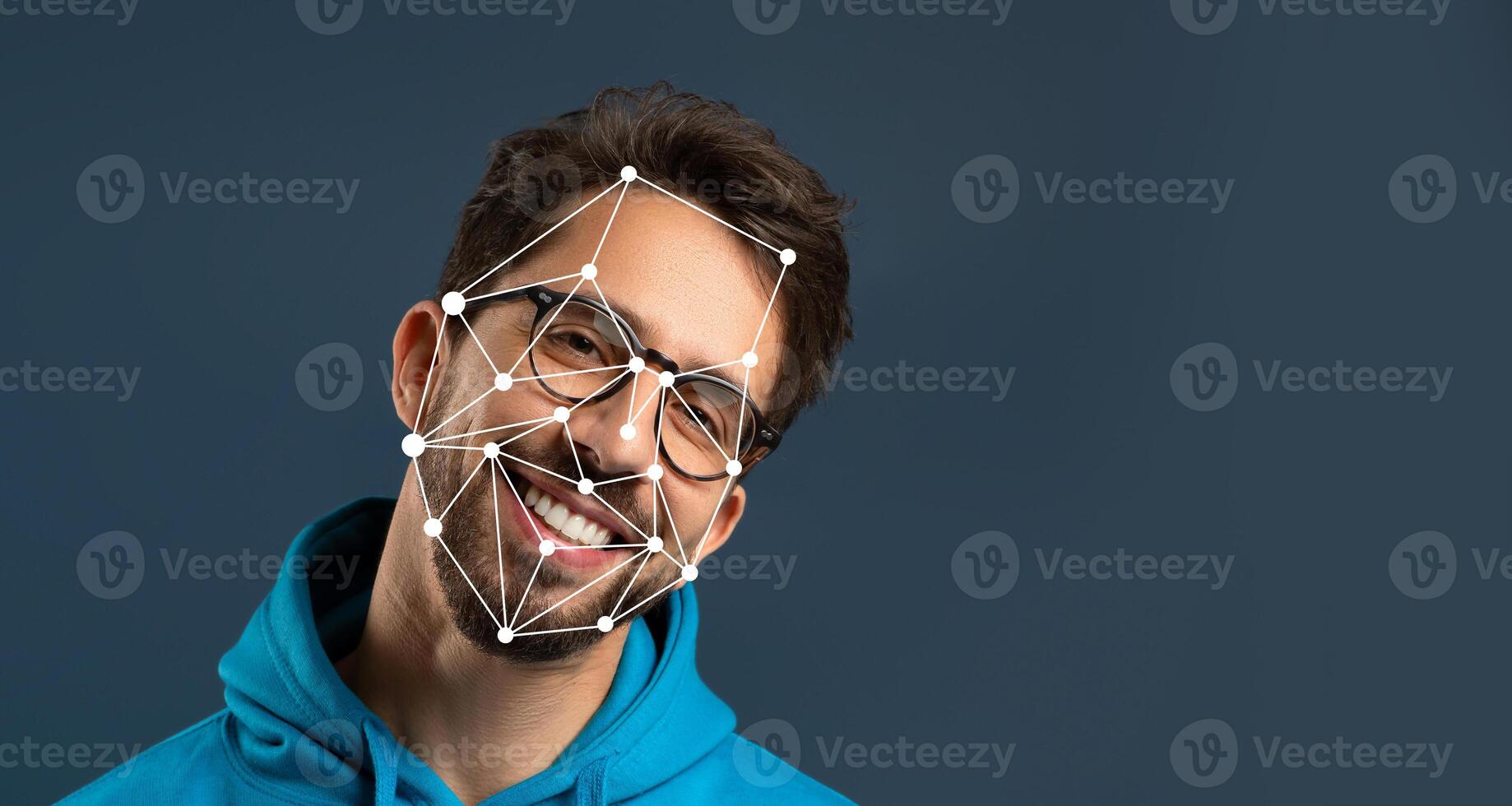 A young man with glasses smiles brightly while wearing a hoodie. The background is dark, highlighting facial recognition markers on his face, illustrating advances in technology. photo