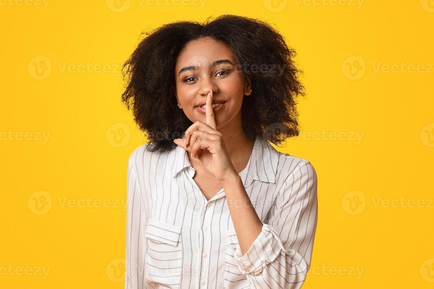 Keep silence. Cheerful afro girl showing shhh sign with finger near lips, standing over yellow background and looking at camera photo
