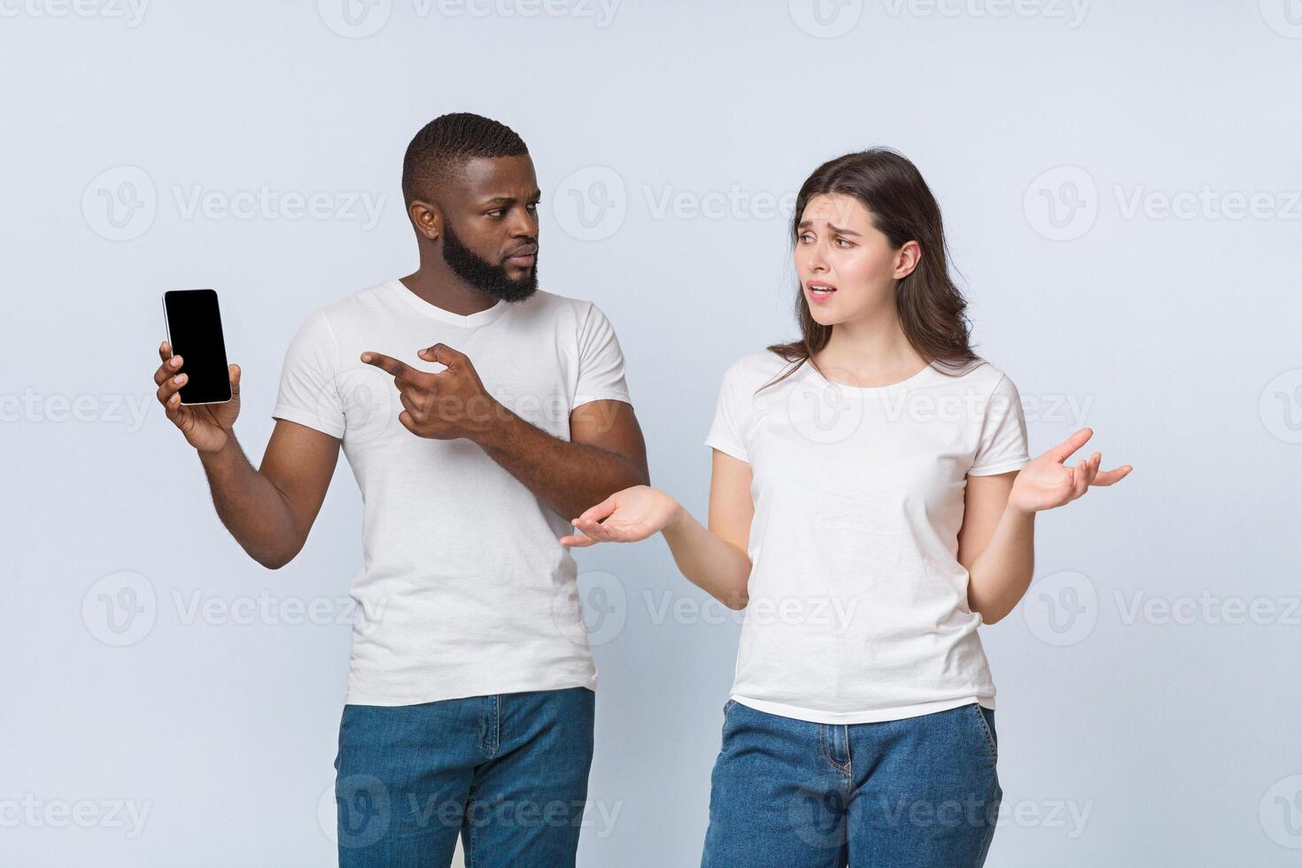 Jealous black boyfriend holding smartphone and asking explanation from his cheating girlfriend, standing together over light background in studio photo