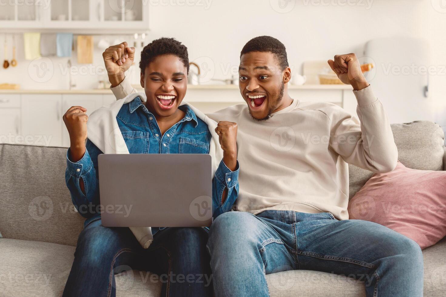 Win. Portrait of excited black couple looking at laptop computer, celebrating success shaking clenched fists sitting on the sofa at home, reading great news, screaming yes, buying something with sale photo