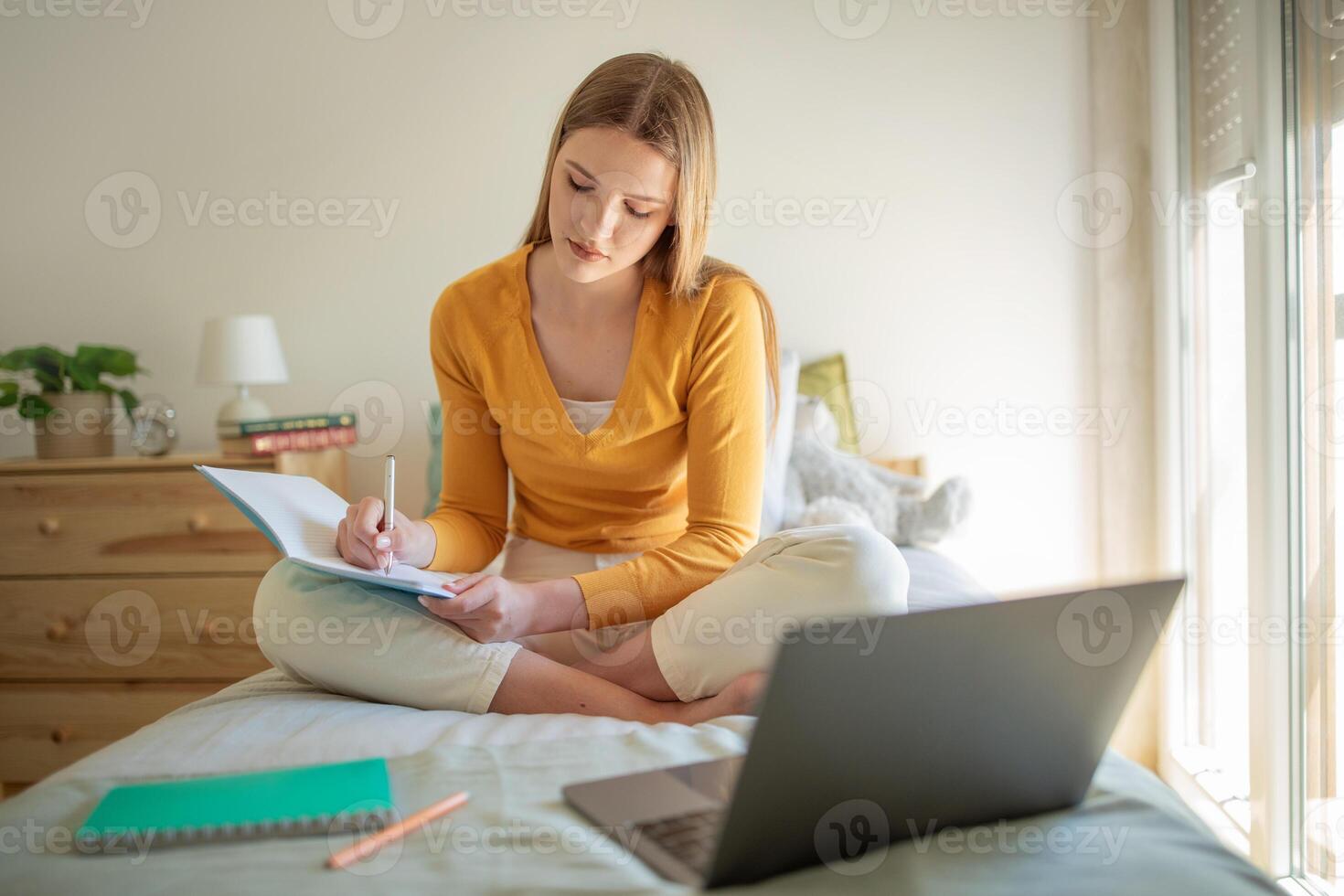 A teen girl sits cross-legged on her bed, focused on writing notes in a notebook. A laptop rests nearby on the bed, and sunlight filters through the room, creating a warm atmosphere. photo