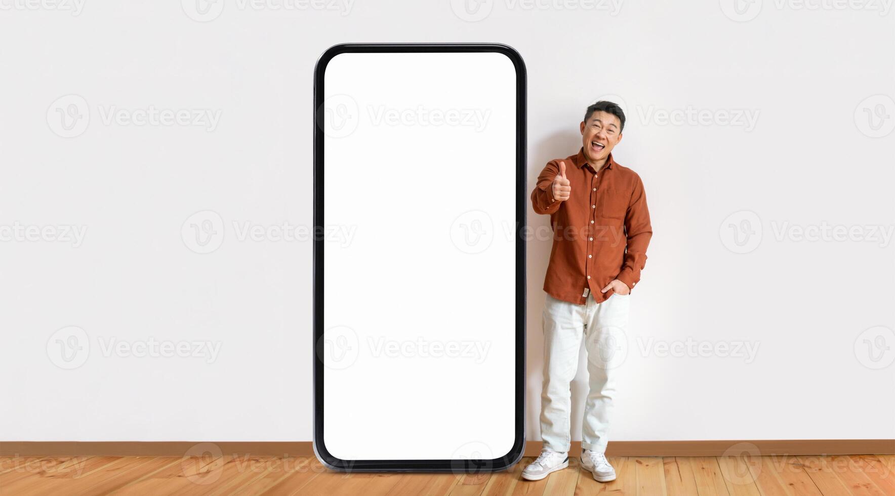 Asian man stands beside a giant blank smartphone, smiling and giving a thumbs up, indicating excitement or approval in a bright and minimalist setting with wooden flooring. photo