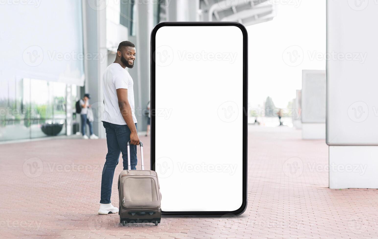A young black man smiles at the camera while standing beside a large blank screen. He has a suitcase by his side, suggesting he is ready for travel at a busy transportation location. photo