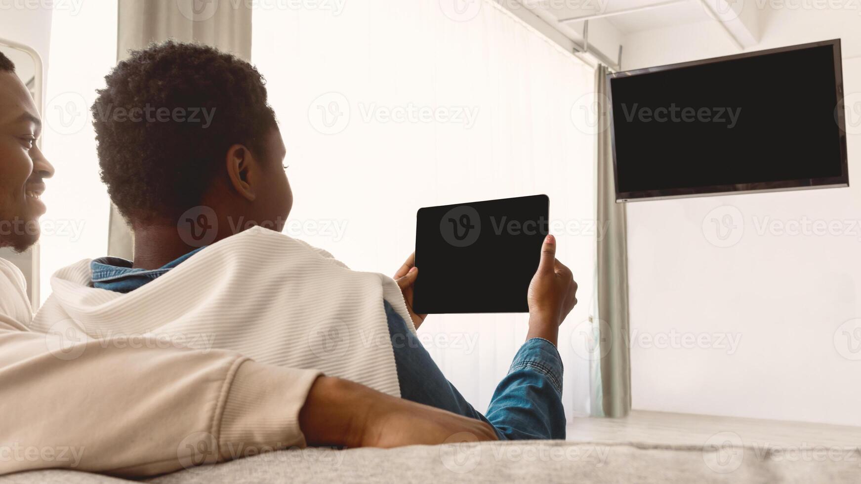 African American couple holding and using digital tablet, watching or movie, sitting on couch looking at black blank screen for mock up template, selective focus, blurred background photo
