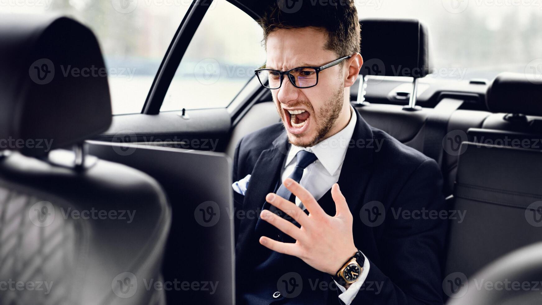 Mad bearded businessman in glasses working on laptop, sitting in backseat of luxury car, gesturing angrily photo