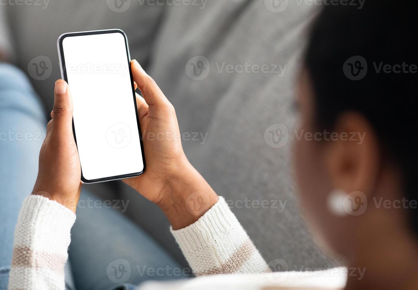 Unrecognizable Black Woman Holding Smartphone With White Screen At Home, Over Shoulder View Of African American Lady Using Blank Mobile Phone While Relaxing On Couch, Mockup Image With Copy Space photo