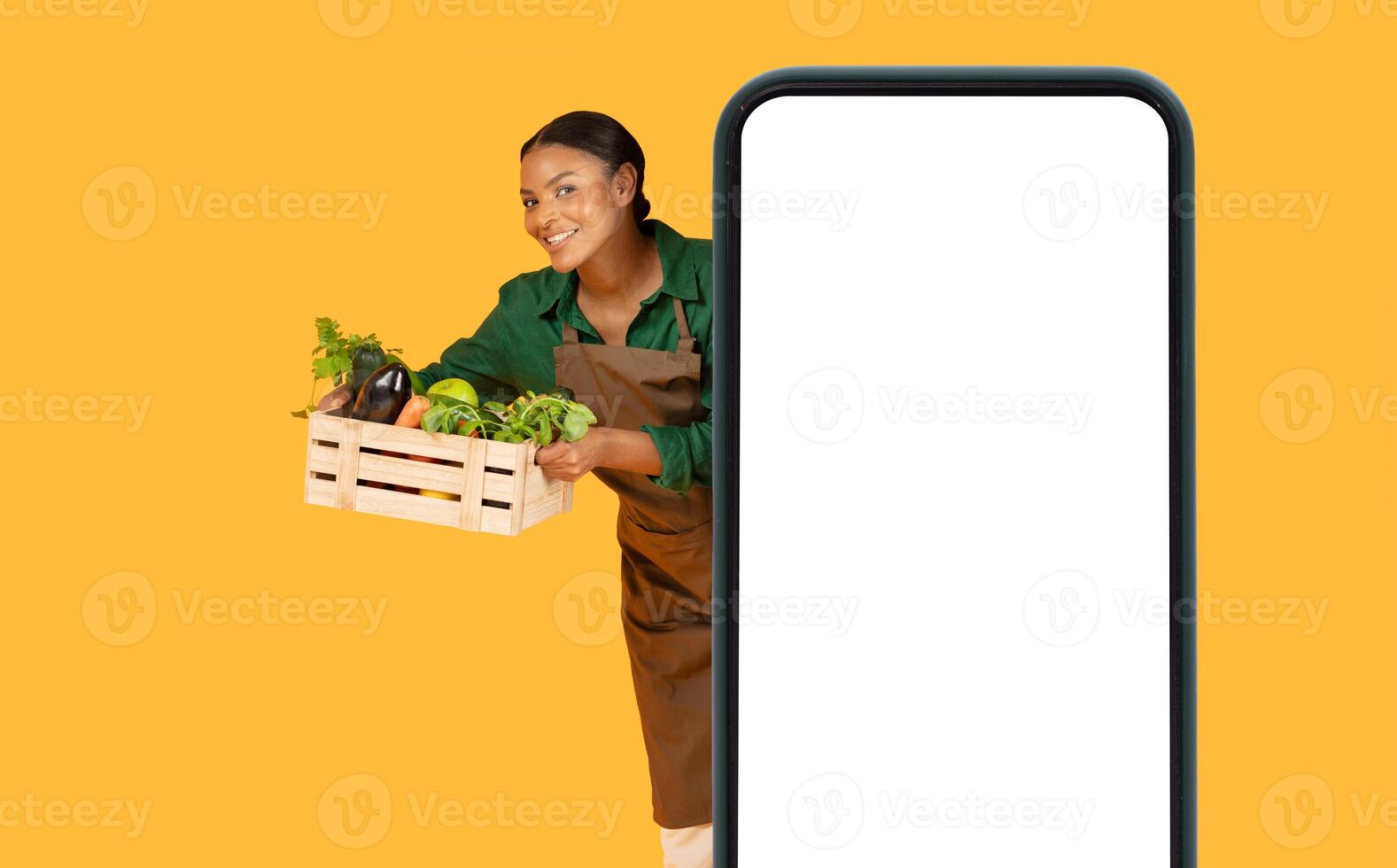 A smiling black woman in an apron stands beside a large smartphone display while holding a wooden crate filled with fresh vegetables. She appears enthusiastic about promoting healthy eating. photo