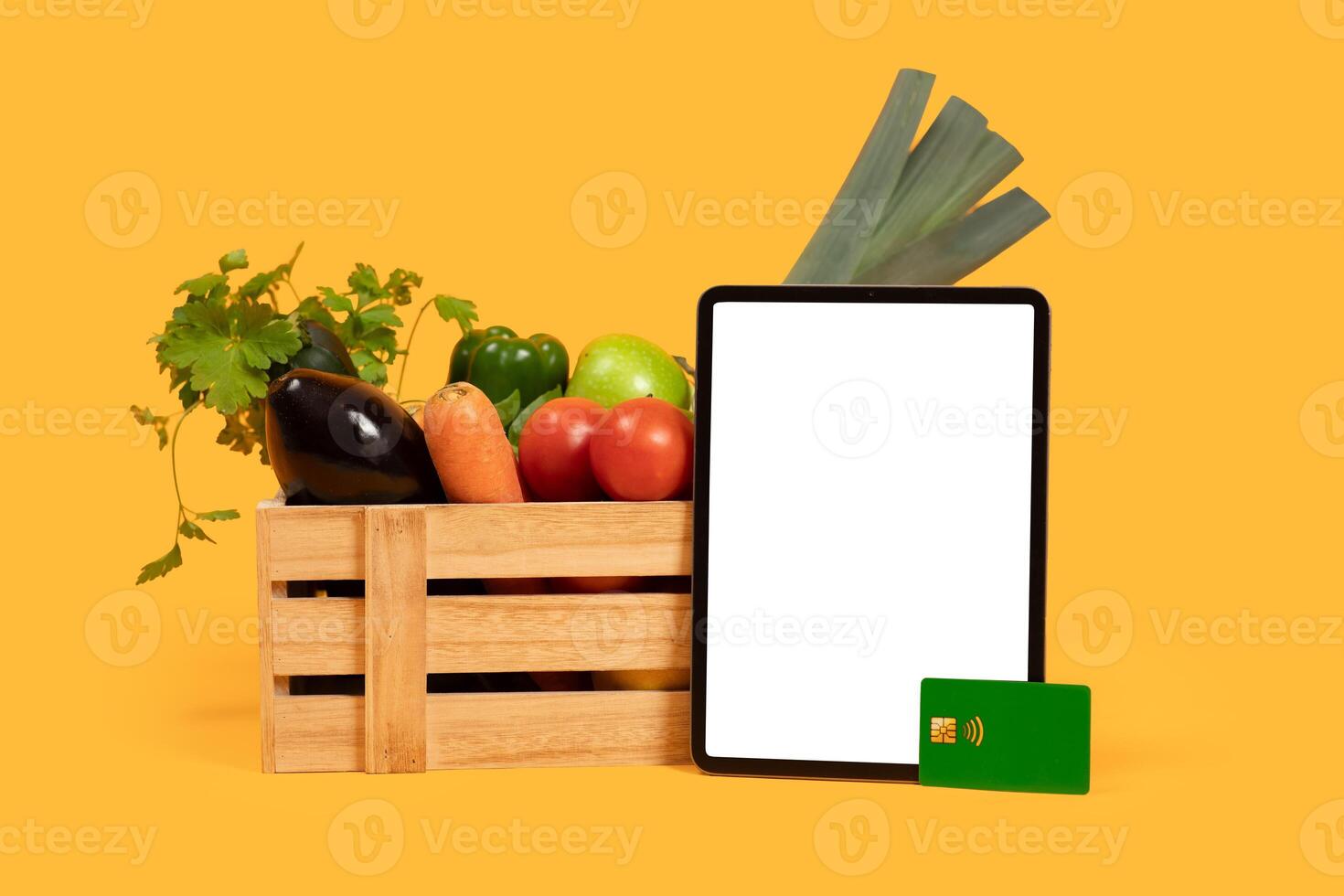 A variety of fresh vegetables, including eggplant, carrots, and peppers, sit in a wooden crate alongside a tablet with blank screen and payment card on a vibrant yellow background. photo