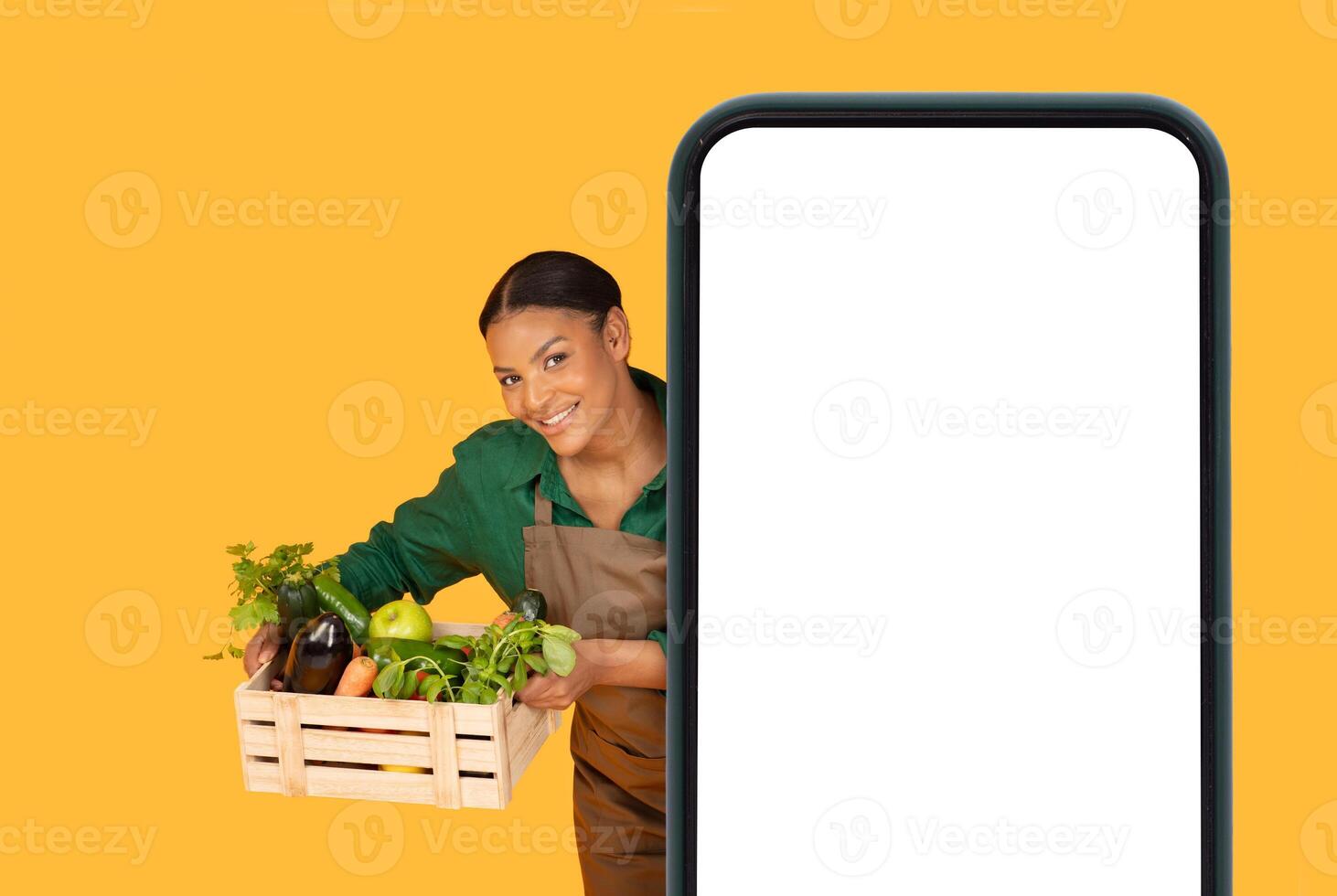 A cheerful young black woman stands with a wooden crate filled with vibrant vegetables, smiling confidently against a bright yellow background while holding an empty smartphone placeholder. photo