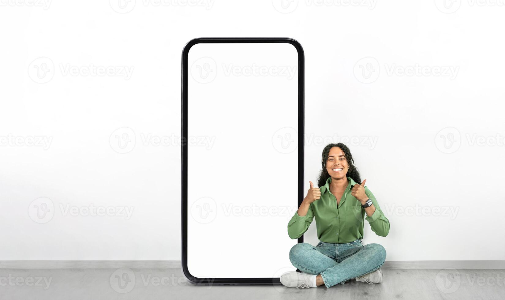 A woman is sitting on the floor, giving a thumbs up with a joyful expression beside a large, empty smartphone frame against a white wall in a bright room. photo