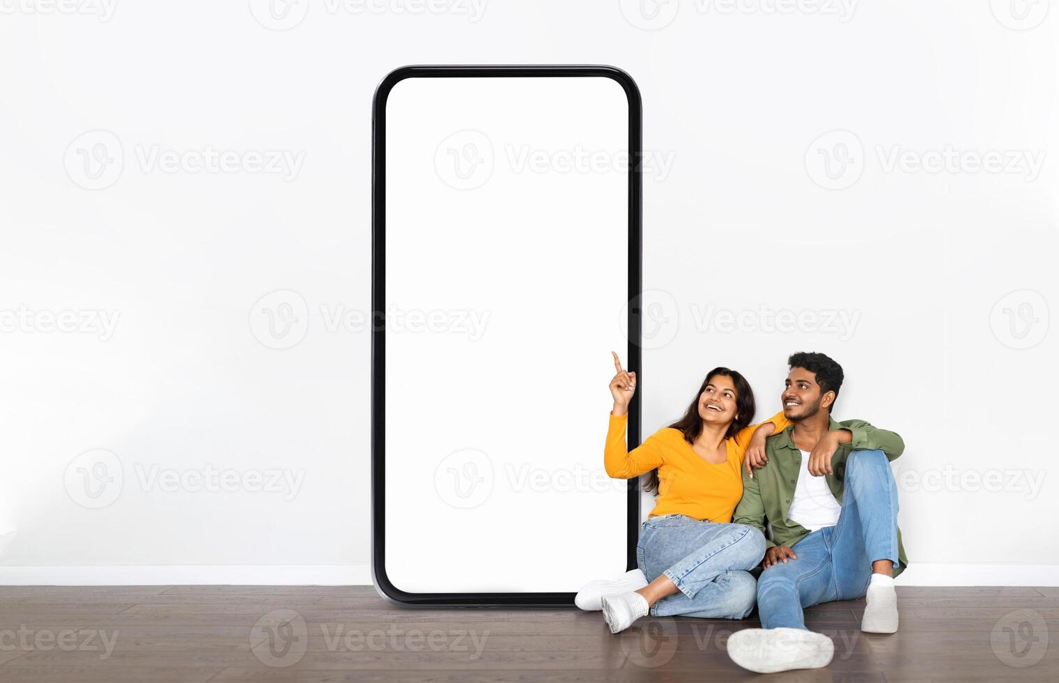 A young Indian couple sits casually on the floor, smiling and sharing a moment together near a large blank phone display. The setting is relaxed, with a simple background emphasizing their joy. photo