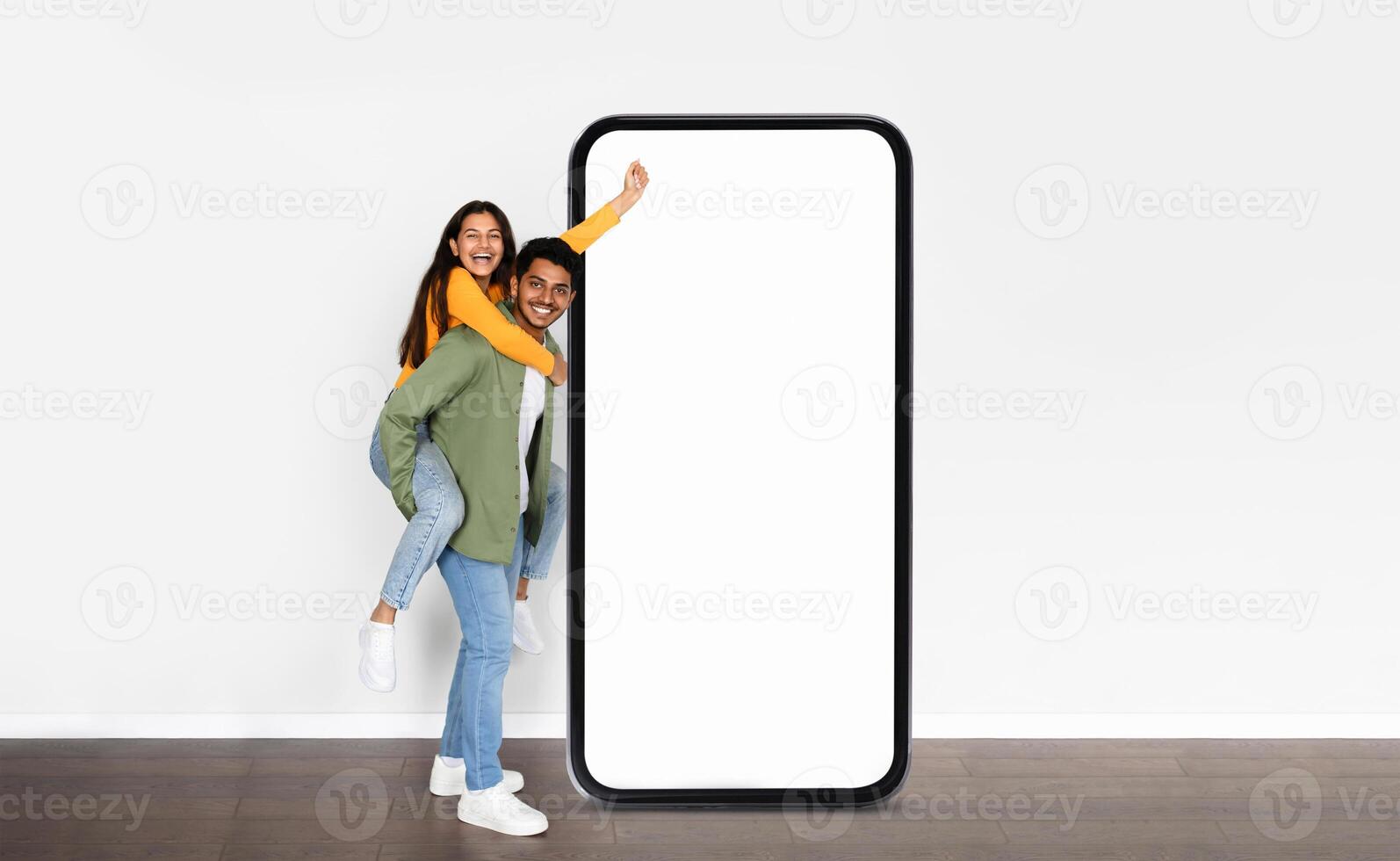 A cheerful Indian couple enjoys a playful moment in a bright space, with one partner giving a piggyback ride while standing next to an oversized smartphone photo