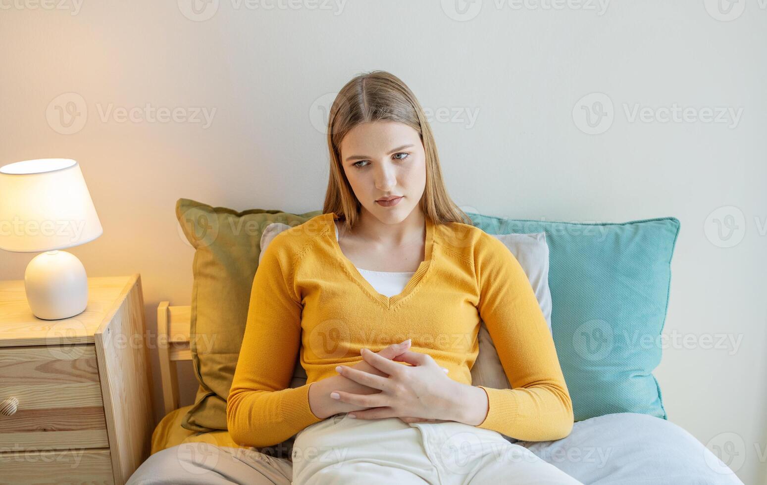 A teen girl is sitting on her bed with a worried expression, wearing a yellow sweater. The soft lighting and decor create a calm yet introspective atmosphere as she thinks quietly. photo