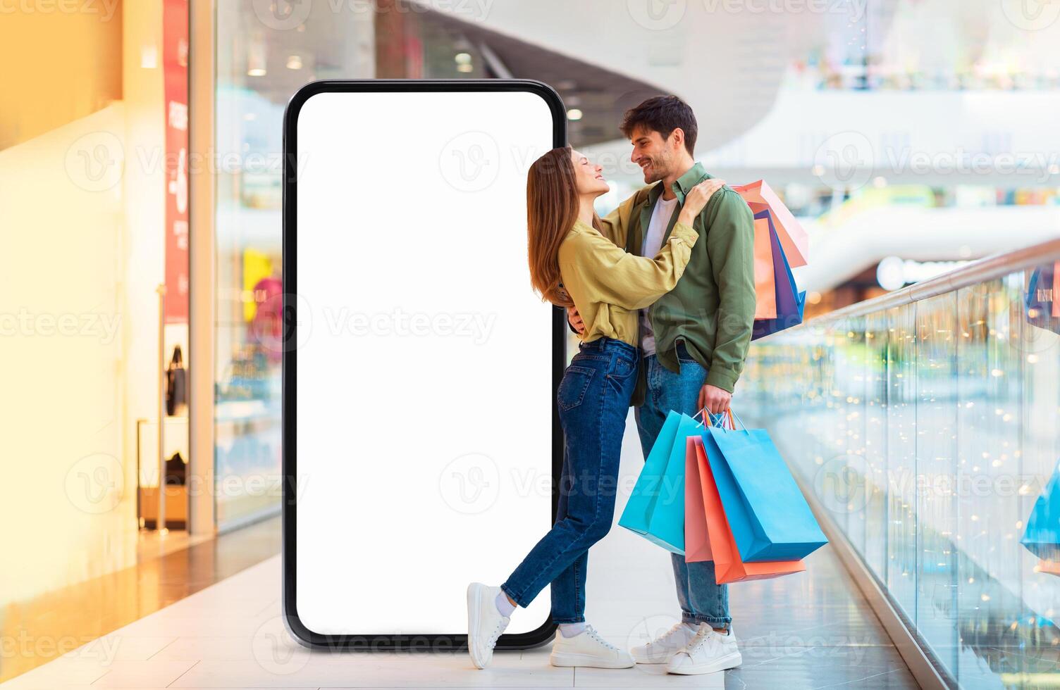 A young couple stands in a bustling mall, smiling at each other while holding colorful shopping bags. They are surrounded by retail displays and shoppers, creating a lively atmosphere. photo