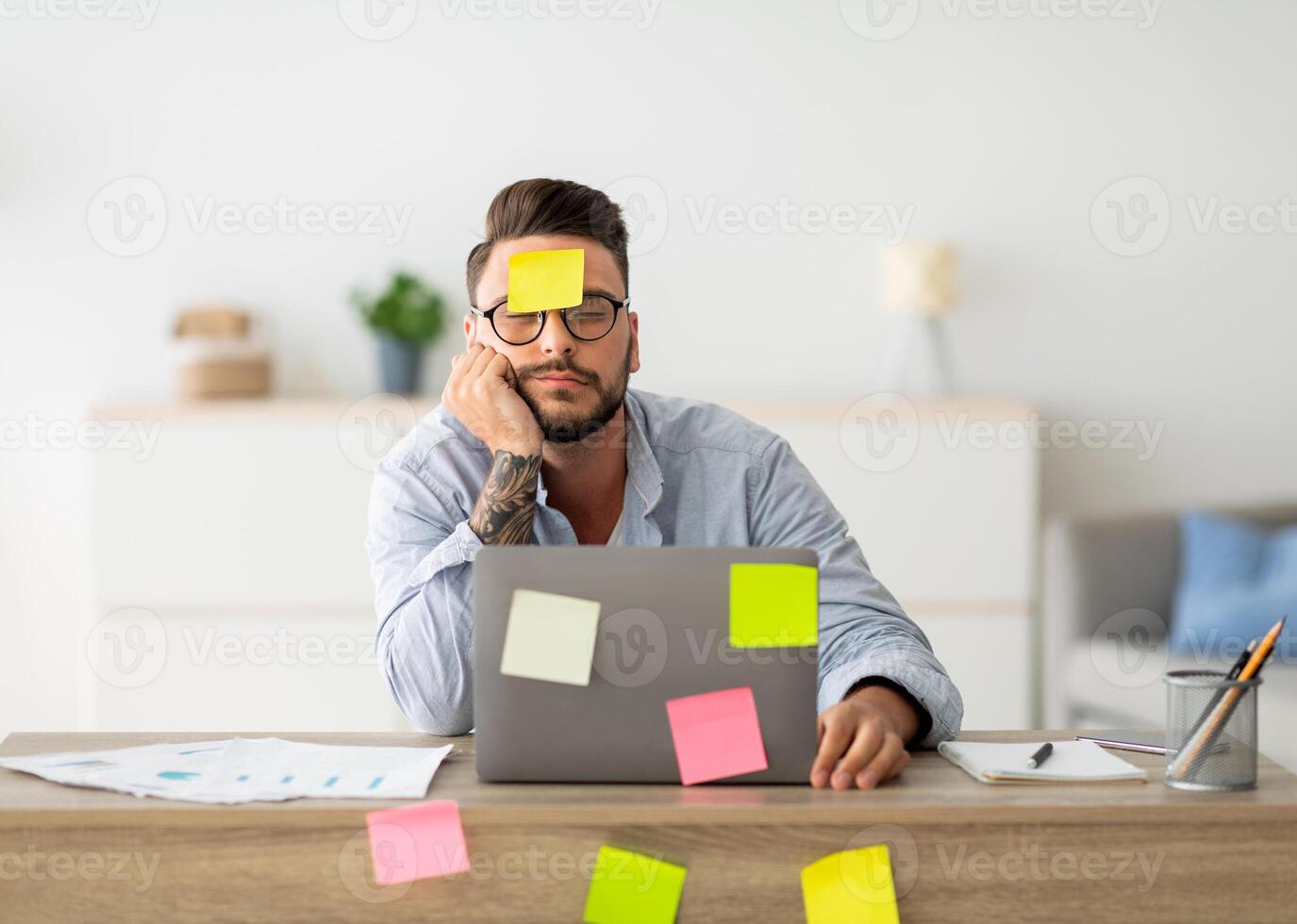 A lot of work concept. Tired freelancer man sleeping at workplace with stickers on foehead and computer, working on laptop at home office, sitting at table in living room, free space photo
