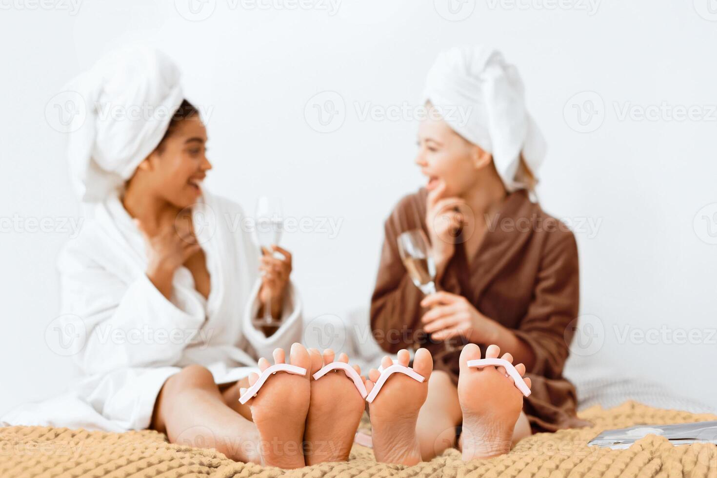 Selective focus on women feet with toe separators on, young multiracial girlfriends having spa day at home, drinking champagne and laughing photo