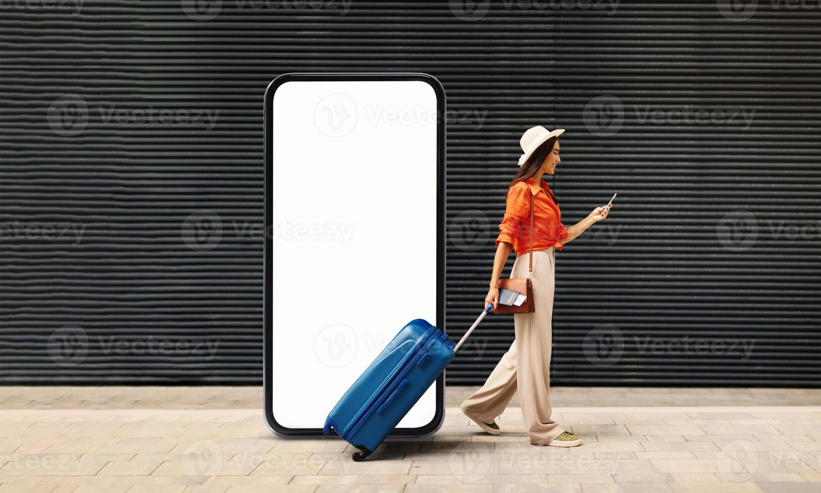 A woman walks with a blue suitcase while checking her smartphone, standing next to a large blank display. This urban setting suggests a busy travel hub during the day. photo