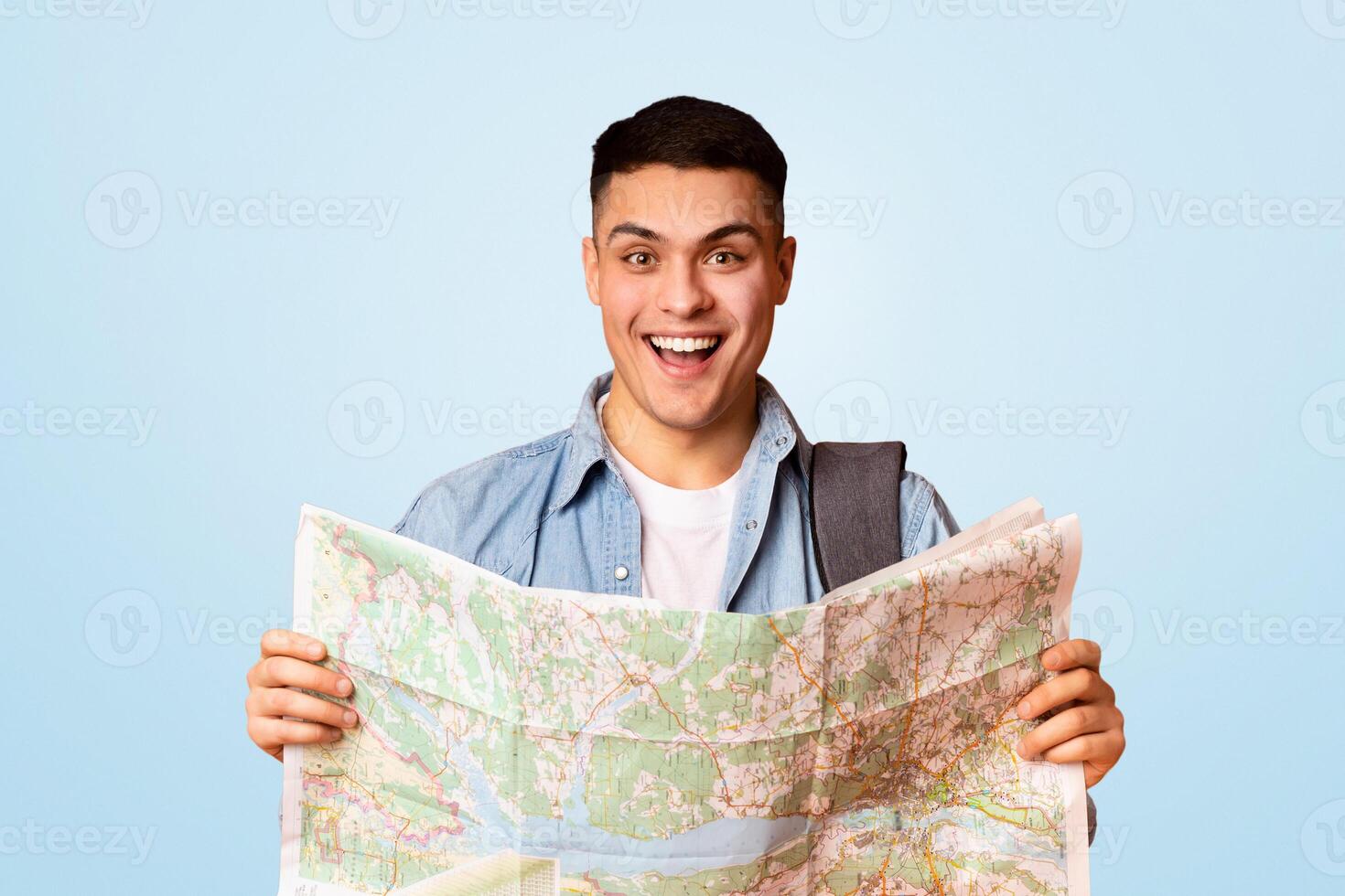 Happy young man traveler with backpack holding city map, orange studio background, sightseeing concept photo