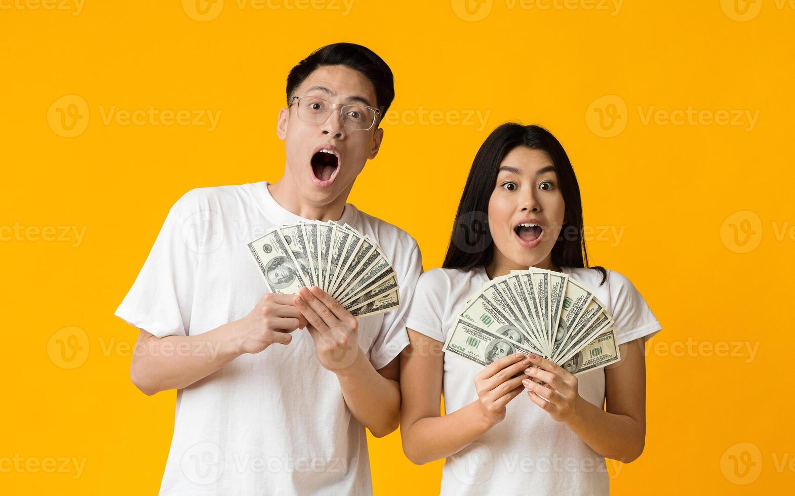 Amazed asian couple holding bunch of money banknotes and celebrating, yellow studio background photo
