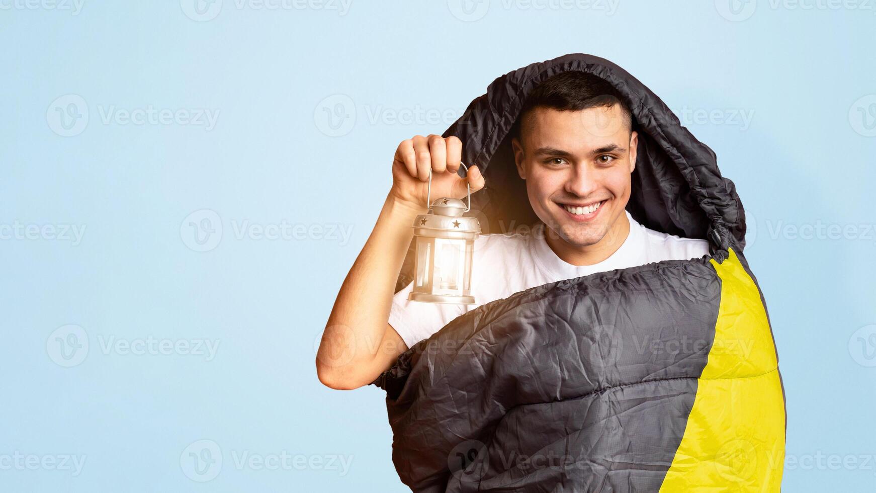 Funny male hiker hiding into sleeping bag, holsing lamp, orange background, camping concept photo