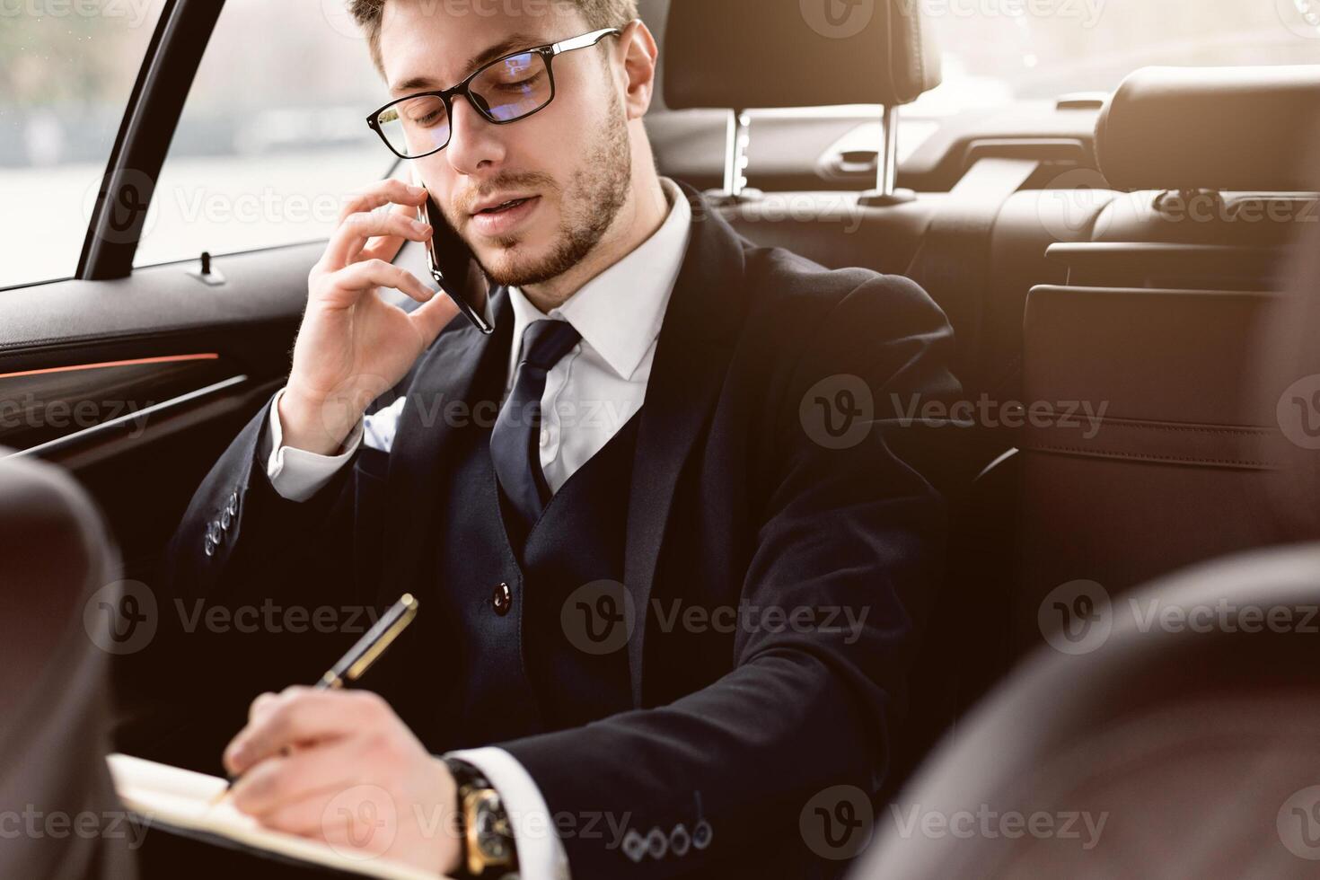Always In Touch Concept. Young manager in glasses writing in notebook talking on smart phone sitting in back seat of car photo