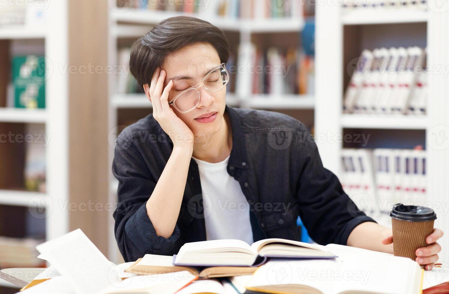 Exhaustion Concept. Sleepy korean guy in glasses napping in library, leaning on elbow, holding cup of coffee photo