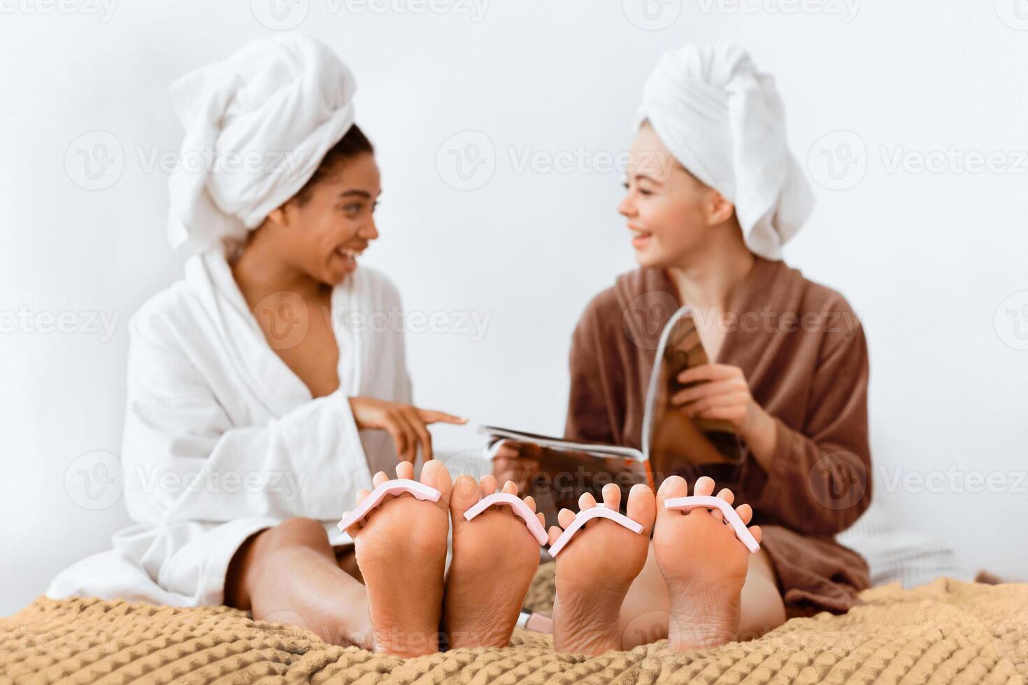 Multiethnic girlfriends in bathrobes sitting on bed and making pedicure at home, reading magazine and talking photo