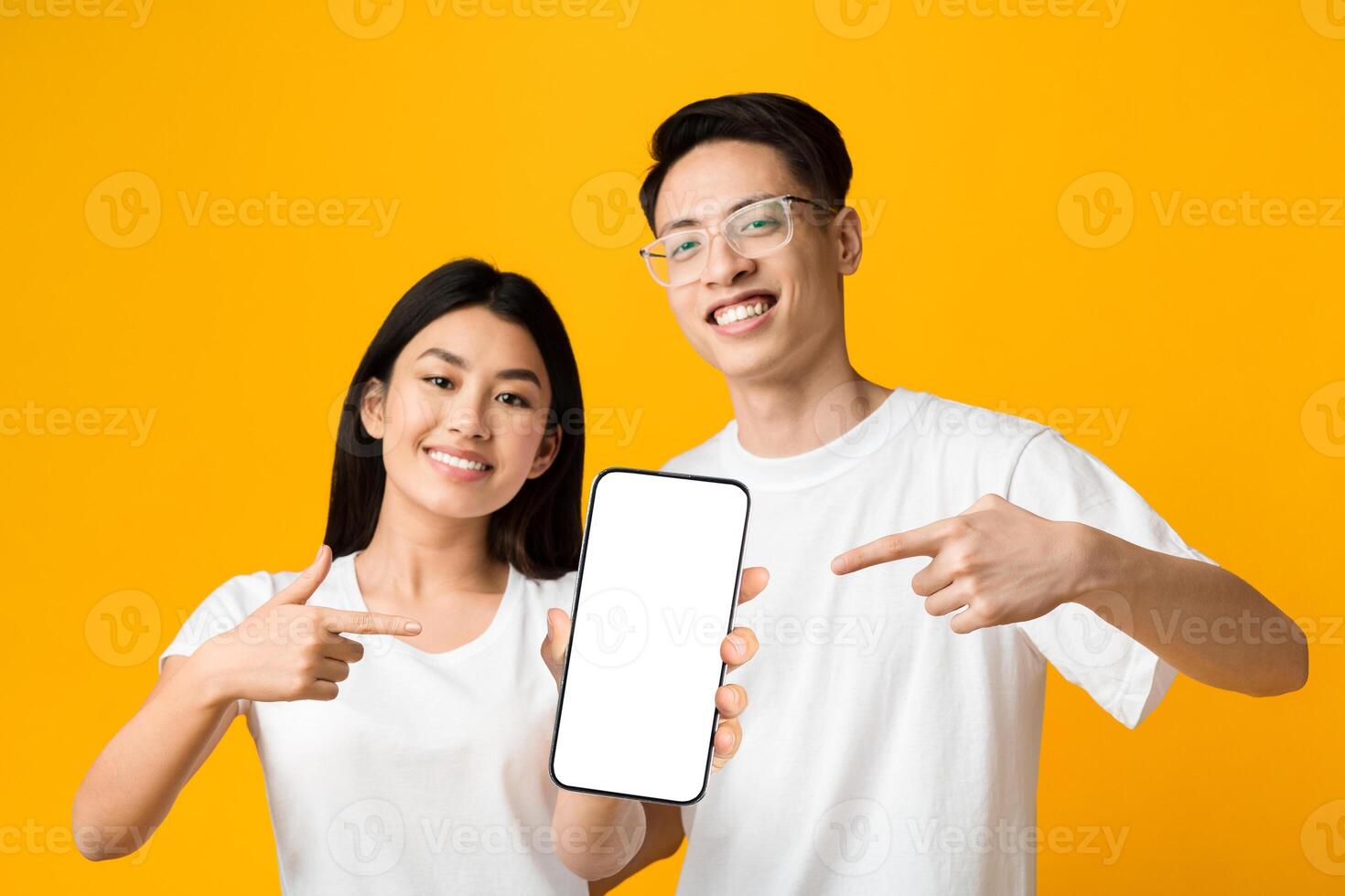 We like this app. Cheerful asian couple holding and pointing at smartphone with blank screen, yellow background photo