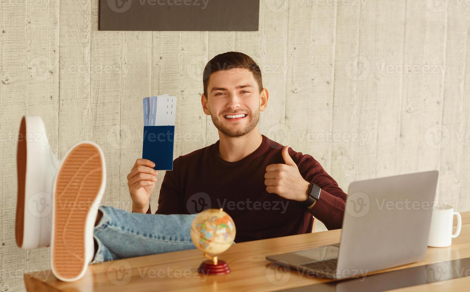Planning Vacation. Happy Guy Holding Flight Tickets Gesturing Thumbs Up Sitting At Laptop In Office. photo