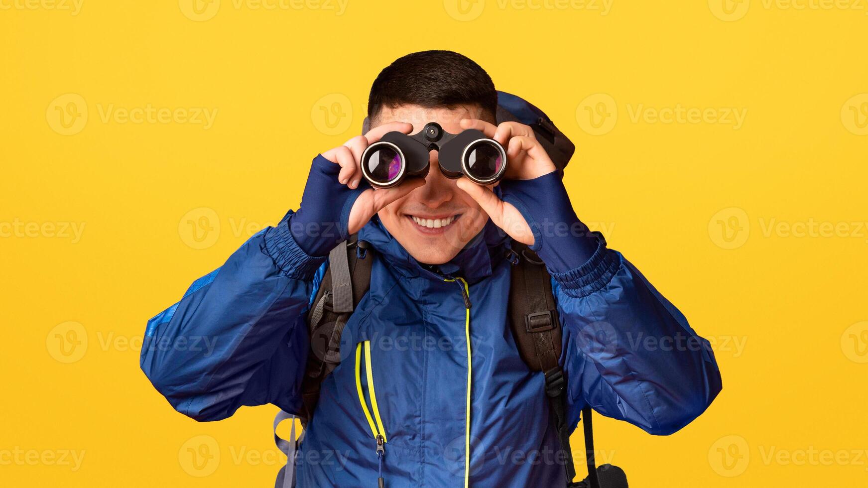 Portrait of smiling young guy hicker using modern binoculars, orange background photo