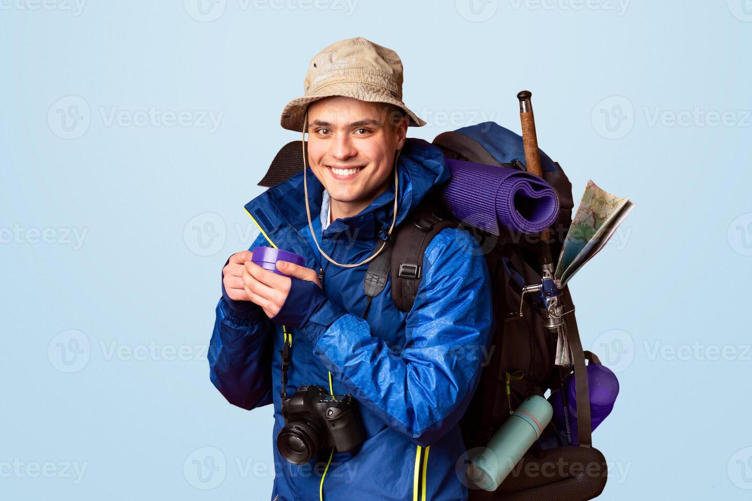Fully equipped young cheerful backpacker drinking hot tea or coffee, wearing jacket, hat, backpack full of diverse tools, orange studio background photo