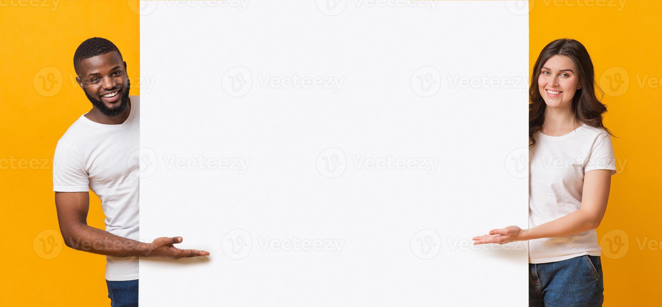 Smiling Multiracial Couple Holding And Pointing At White Advertisement Board, Standing Together Over Yellow Background, Free Place For Your Text photo