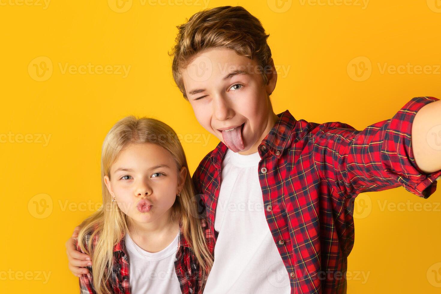 Playful kids taking selfie, little brother and sister making funny faces and fooling together while capturing photo, yellow background, free space photo
