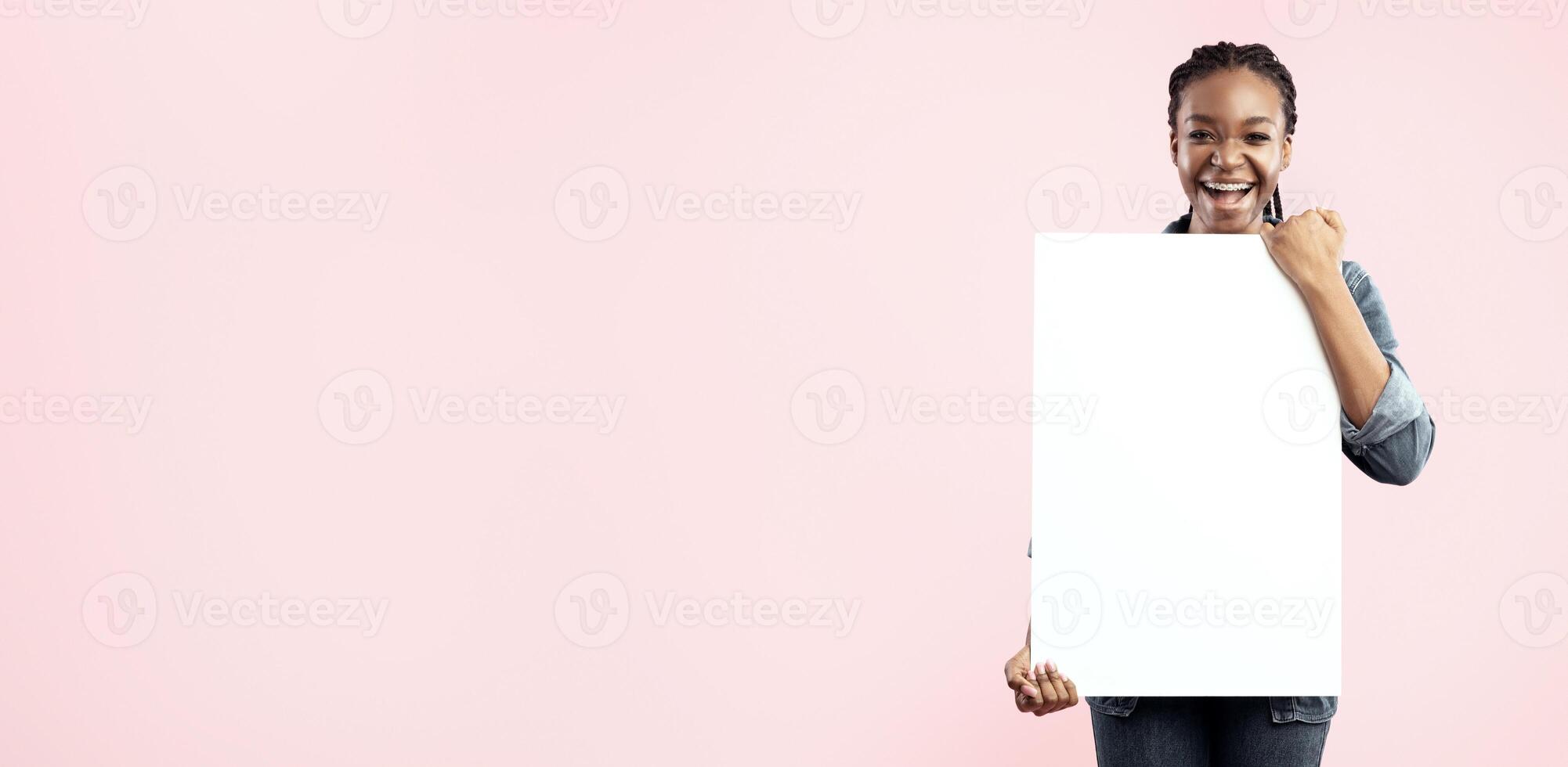 Joyful Black Lady Holding Empty White Paper Board Advertising Your Text Posing In Studio On Pink Background. Mockup, Free Space photo