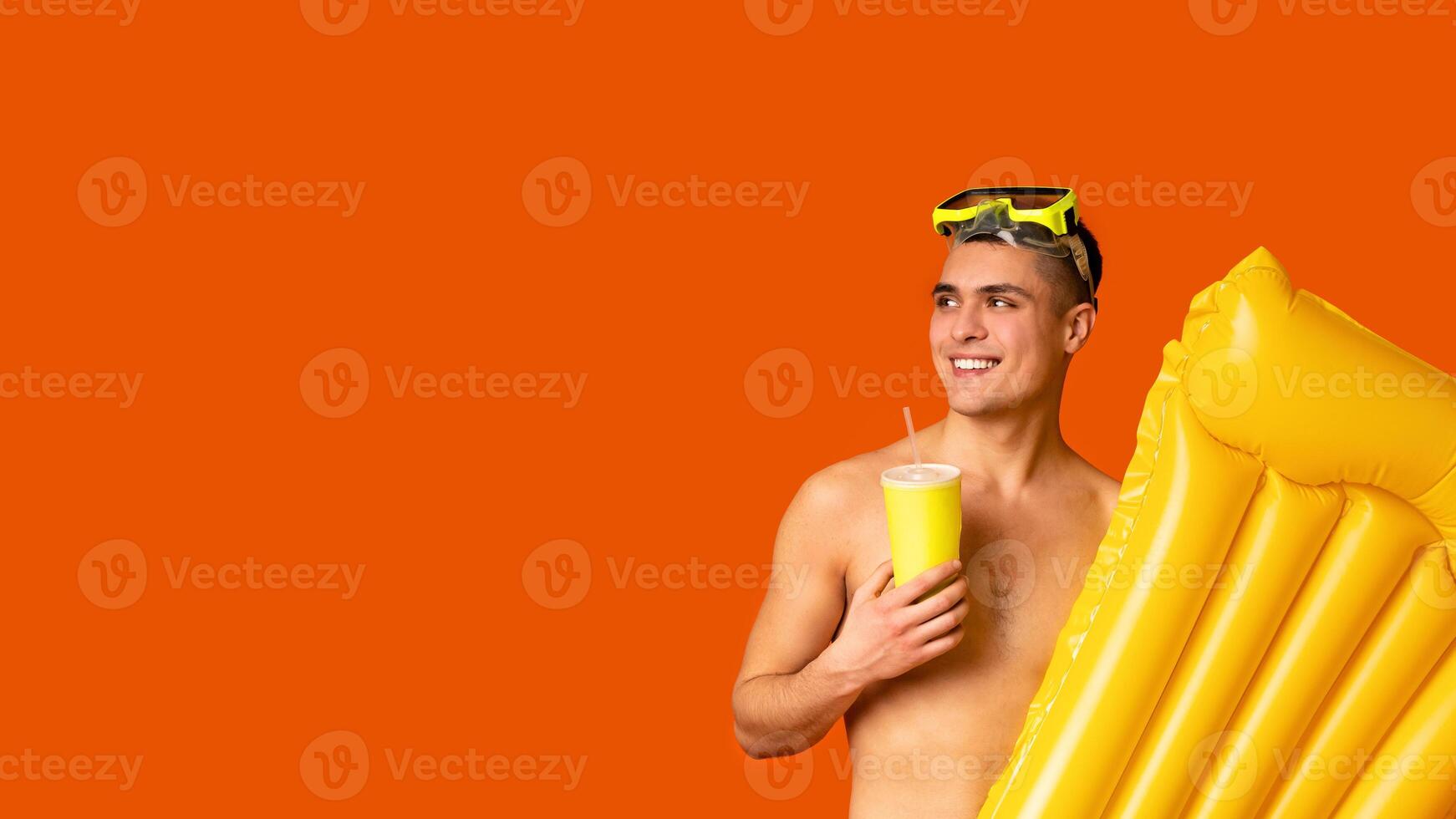 Summer vacation concept. Happy guy with diving mask and air mattress drinking cocktail over orange studio background, looking at empty space, panorama photo