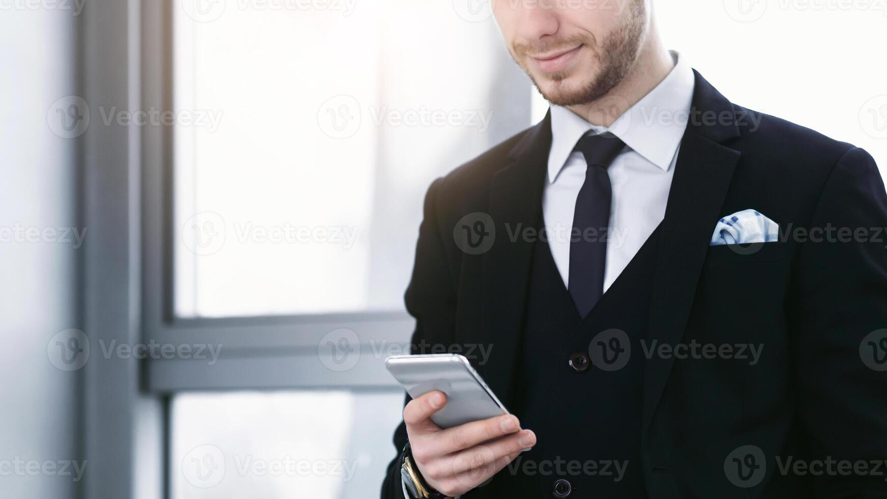 Good News From Colleague. Smiling man holding and looking at phone standing near window at his working place in office photo