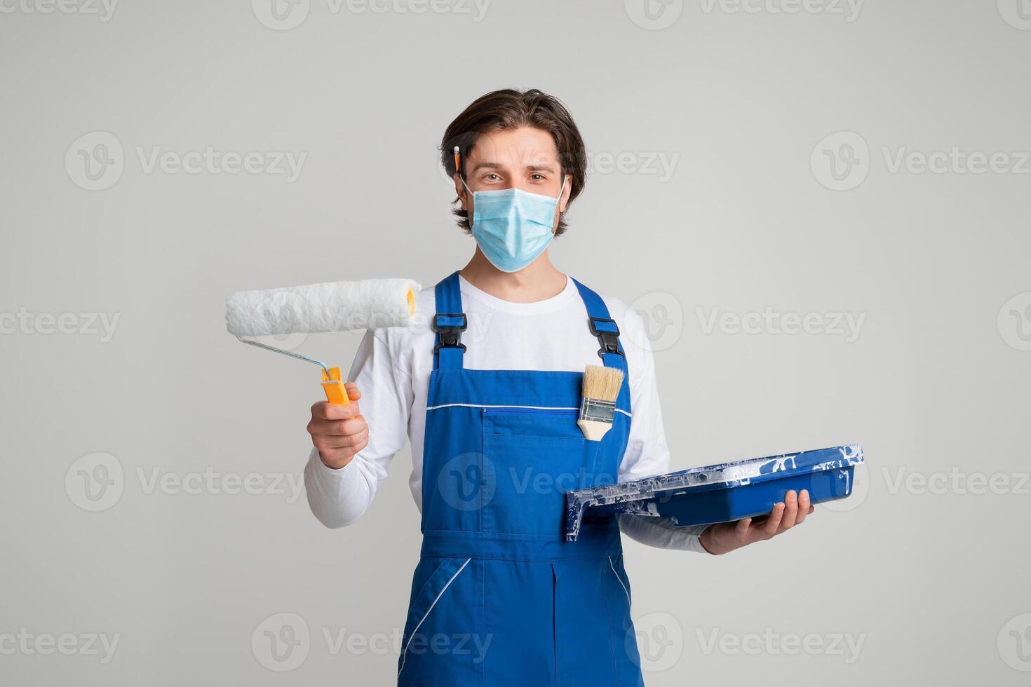 Employment During Coronavirus. Portrait Of Professional Painter In Medical Mask With Paint Roller In Hands On Light Studio Background, Panorama photo