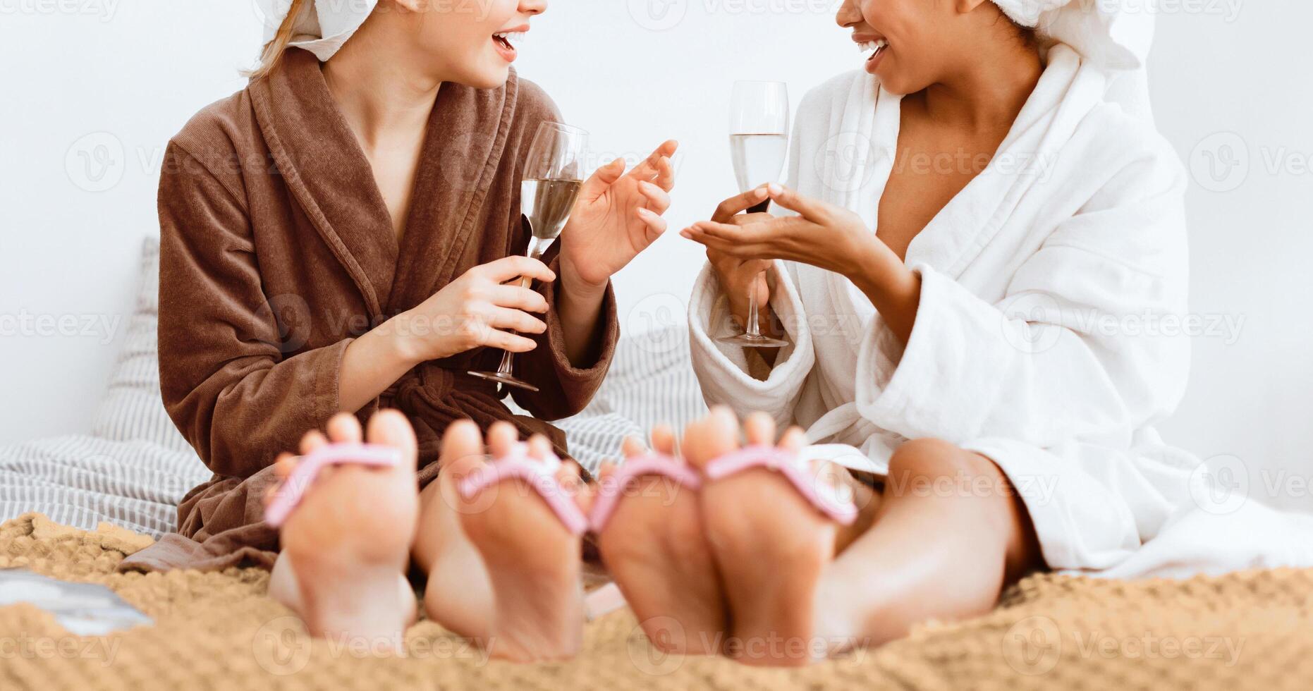 Cropped of two multiethnic girlfriends in bathrobes sitting on bed and drinking champagne, panorama, women friendship concept photo