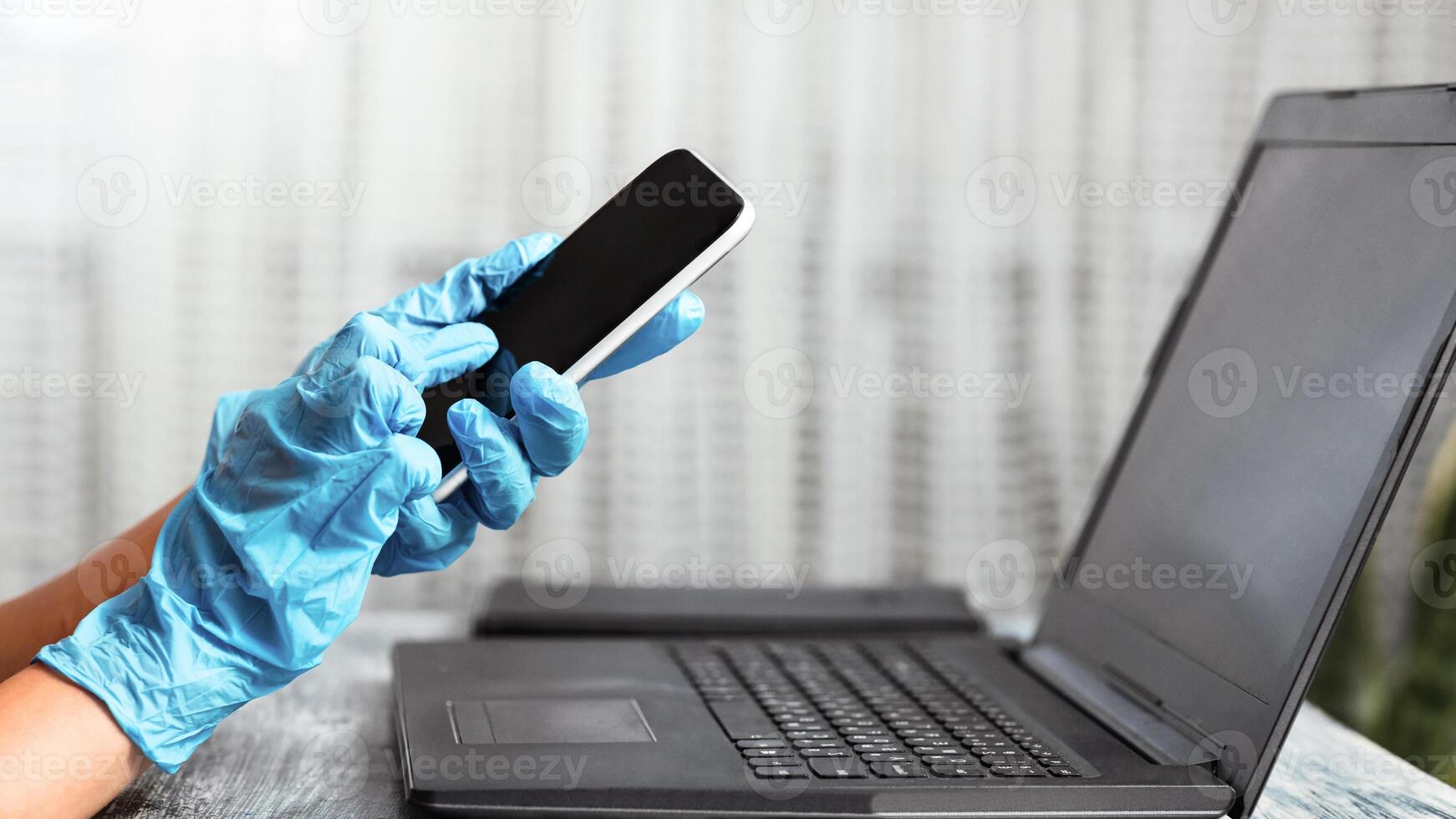Close up on female hands holding smart phone using laptop, wearing protective rubber gloves working during quarantine, mock up photo