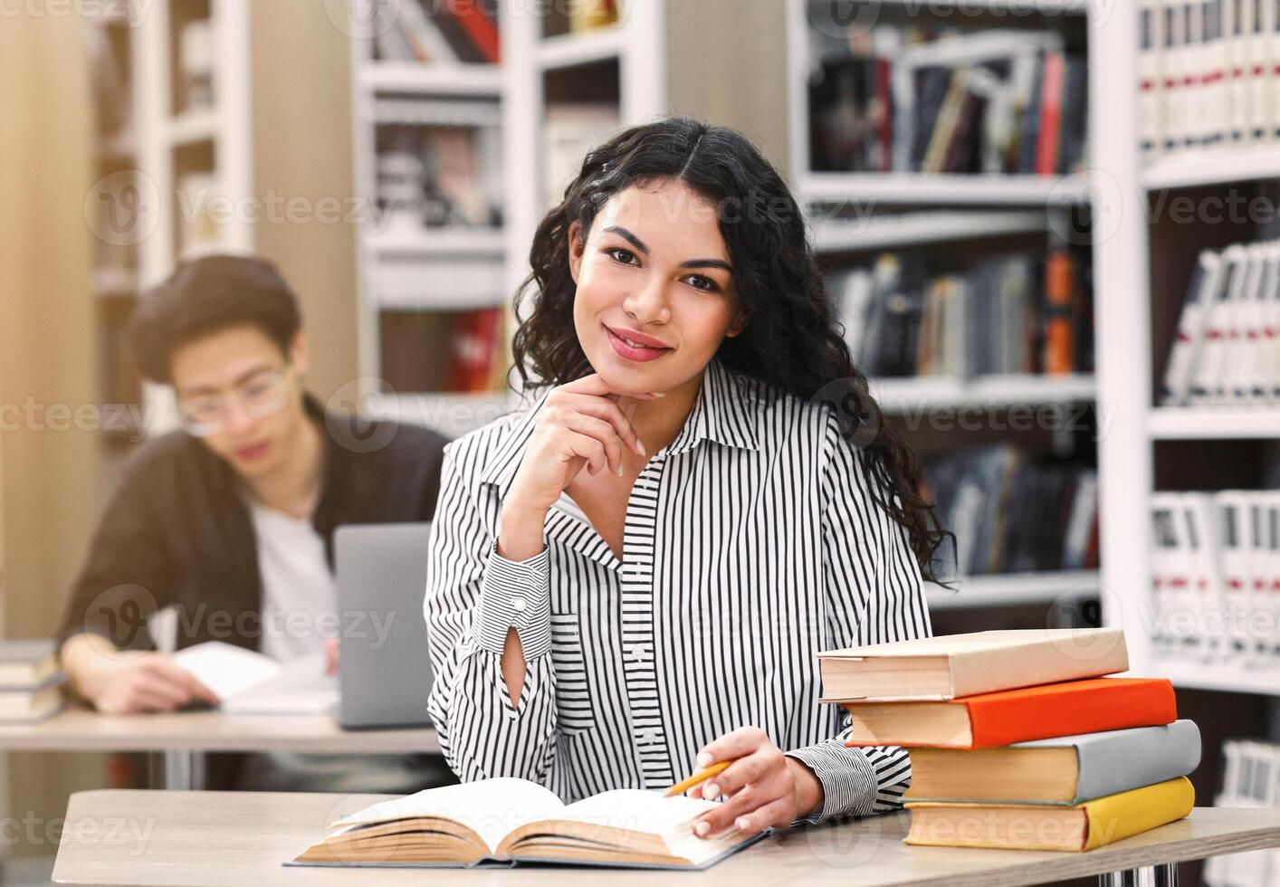 Learning And Educational Concept. Cheerful latin girl sitting at desk in public library, looking at camera, free space photo
