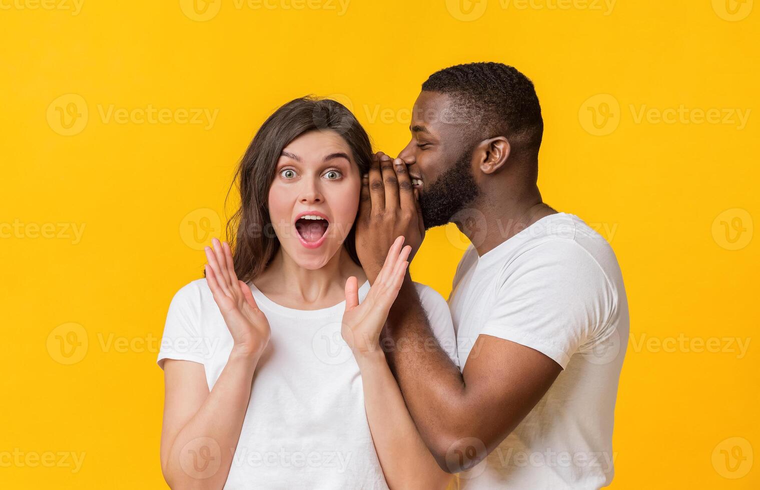 Big secret. Smiling black guy whispering something into his shocked girlfriend's ear, multiracial couple gossiping together, yellow studio background photo