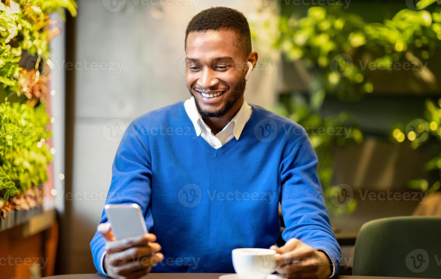 Technology Concept. Front view of happy afro student in headphone using smartphone sitting at modern cafe photo