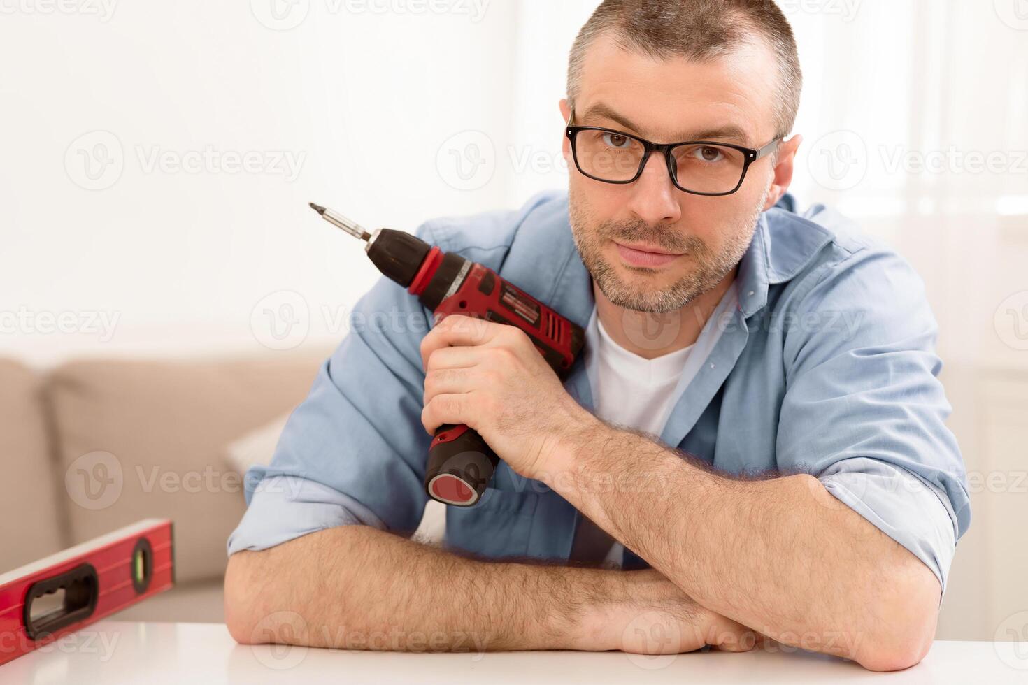 Furniture Assembly. Portrait Of Mature Man Posing Holding Electric Drill And Carpentry Tools Looking At Camera Sitting At Home. Carpenter At Work, Carpentry Service Concept photo